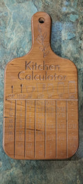 A wooden kitchen calculator cutting board with engraved measurement tables and a hanging hole, shown on a textured stone surface.