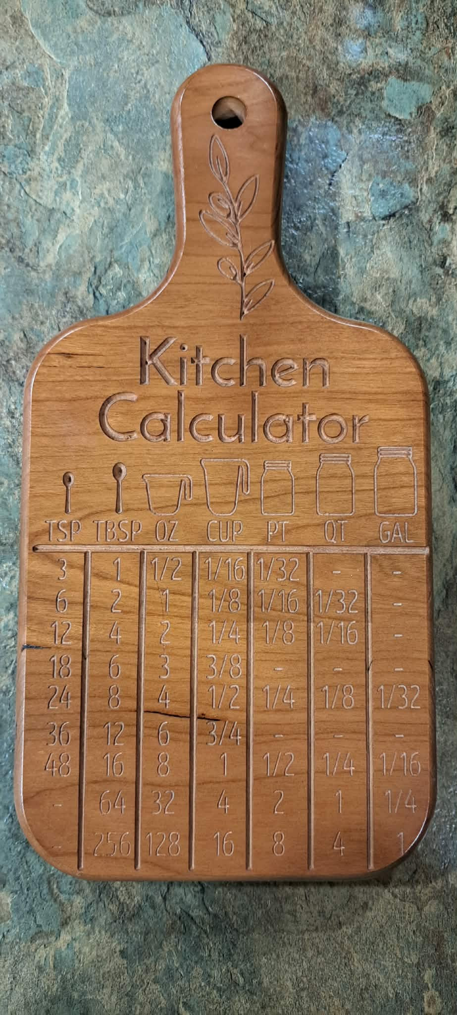 A wooden kitchen calculator cutting board with engraved measurement tables and a hanging hole, shown on a textured stone surface.